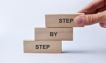 Hand stapelt 3 Holzklötze mit den Aufschriften "Step" "By" "Step"