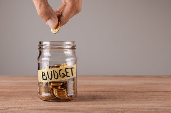 Ein Glasbehälter mit Kleingeld mit der Aufschrift "Budget" steht auf einem Tisch. Eine Hand hält über der Öffnung des Glasbehälters eine Münze zwischen den Fingern.