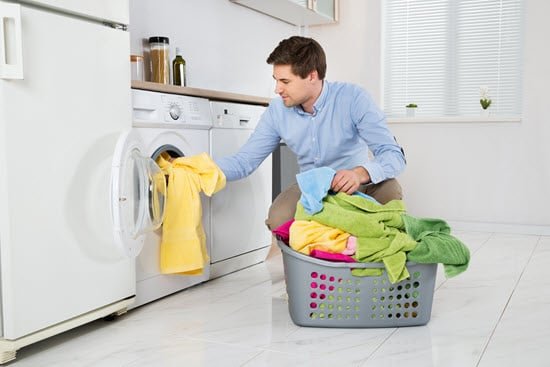 Person mit Wäschekorb voller Handtüchern füllt diese in eine Waschmaschine, welche in der Küche steht. Man sieht noch einen Kühlschrank, eine Spülmaschine und Schränke.
