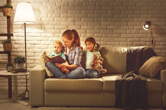 Mutter sitzt mit Kleinkindern auf einem Sofa und liest ein Buch vor. Es scheint bereits abends zu sein und der Bereich ist mit einer Stehlampe und einer Wandleuchte gemütlich beleuchtet.