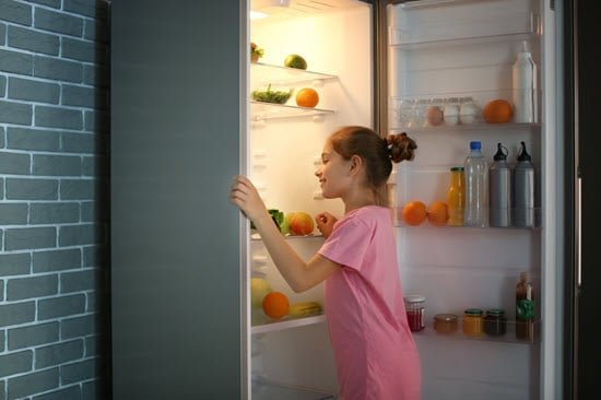 Kind welches am geöffnet Kühlschrank steht und etwas herausnehmen möchte. Man sieht Obst im Kühlschrank und Flaschen in der Tür