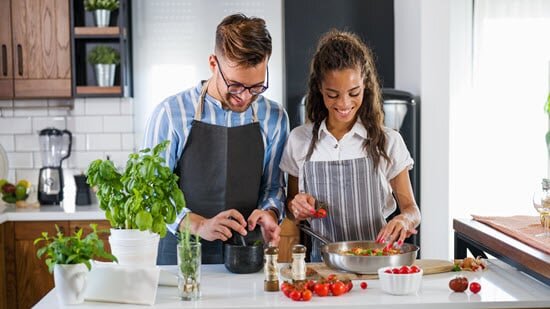 Junges Ehepaar in der Küche am kochen. Man sieht Gewürzpflanzen, Tomaten etc. Eine Person benutzt einen Mörser, die andere eine Pfanne