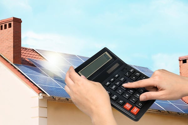 Hände nutzen einen Taschenrechner vor dem Hintergrund eines mit Solarmodulen bedeckten Hausdachs.  | © 2020 New Africa/Shutterstock.